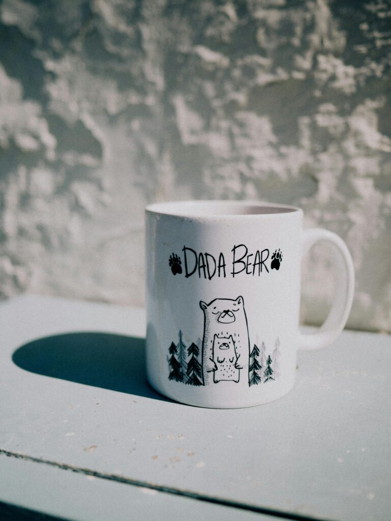 White 'Dada Bear' mug featuring bear doodles on a textured background in sunlight.